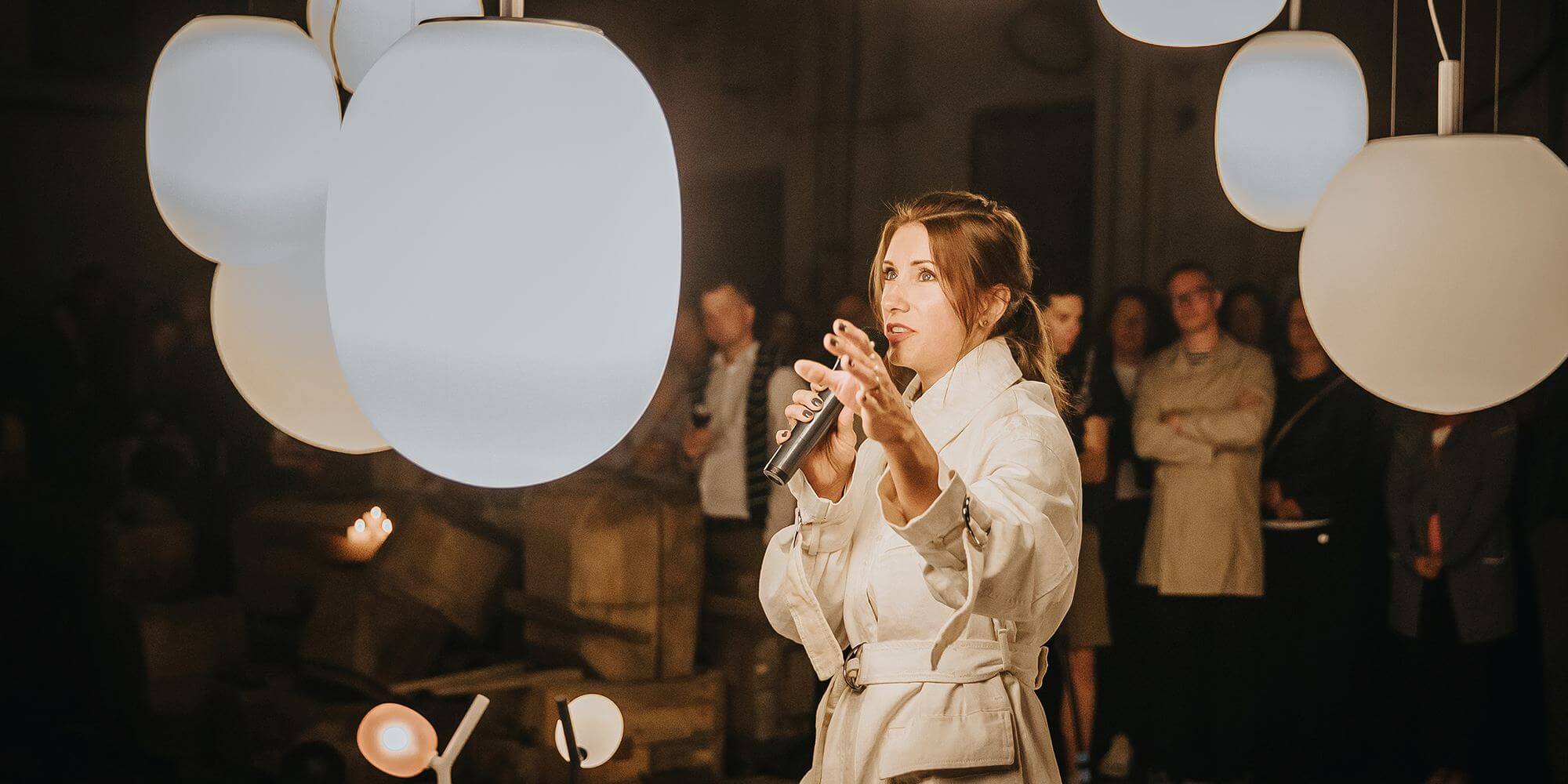 Lucie Koldova speaks into a microphone, framed by sculptural Brokis Sfera pendant lamps and an attentive crowd.
