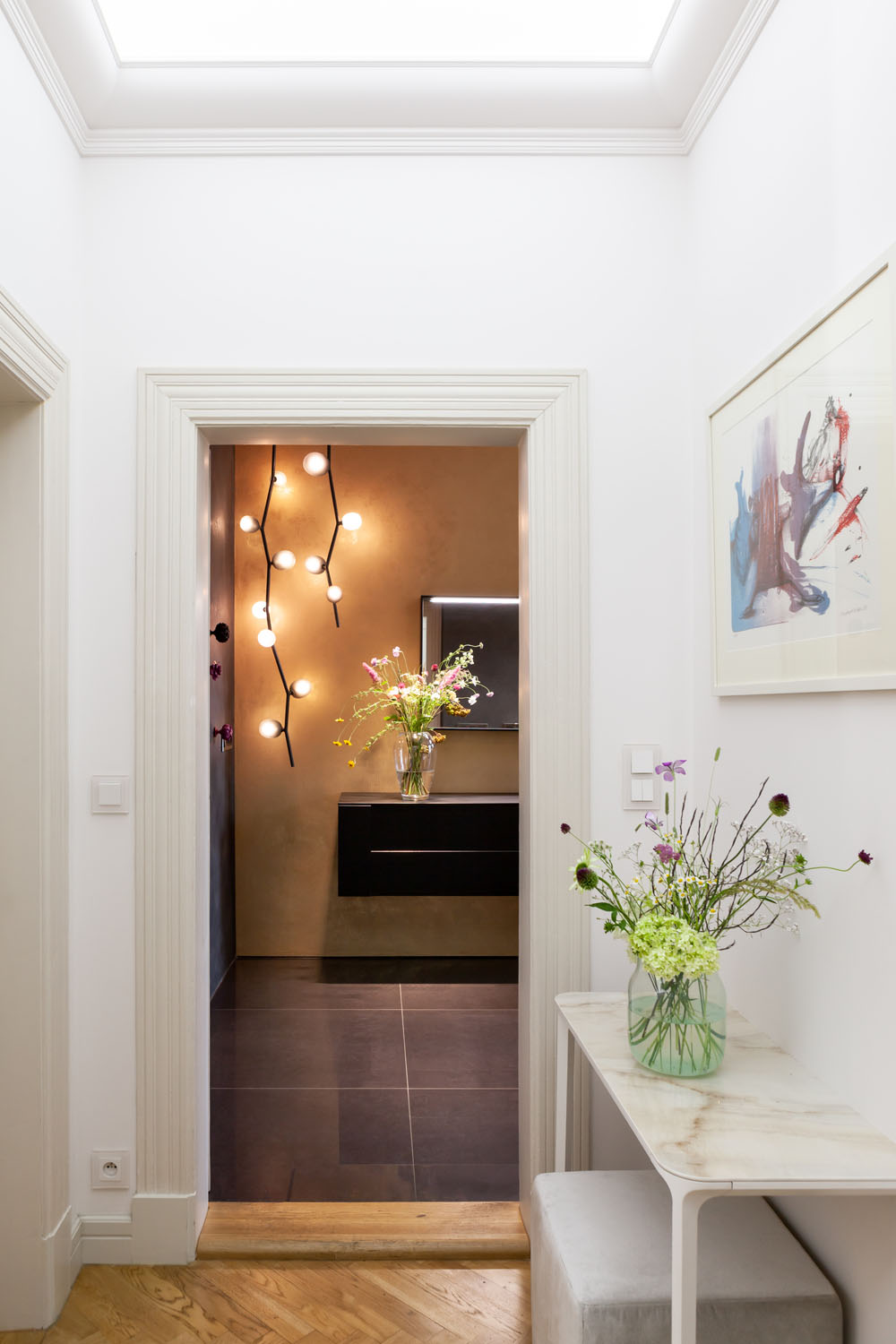 A corridor with white walls, marble console and floral vases leads to a room with Brokis Ivy Vertical pendants.