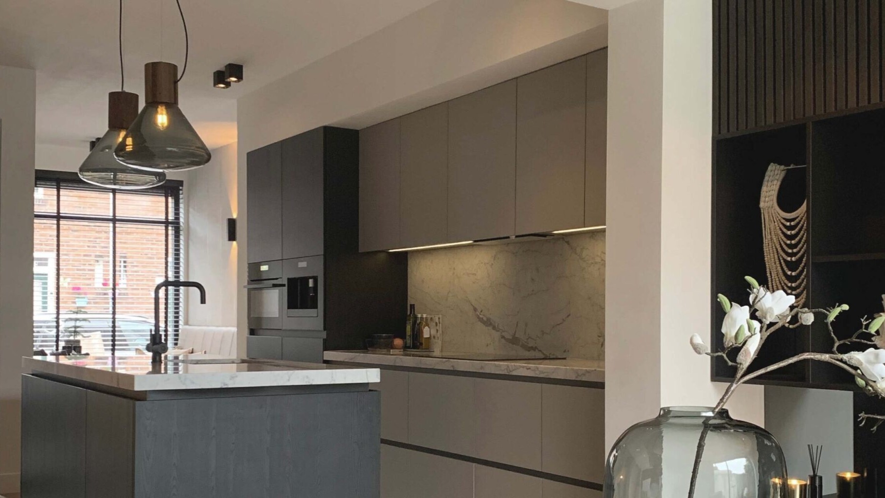 Modern kitchen with grey cabinets, a marble worktop and splashback, pendant lights over an island, large windows letting in natural light, and decorative items including a glass vase with white flowers.