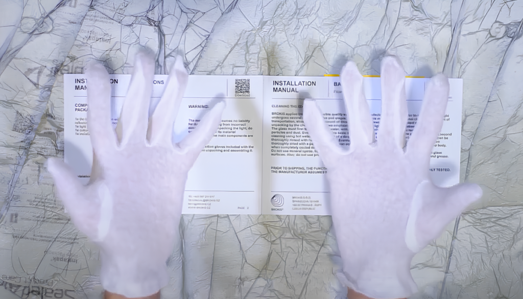 Gloved hands hover over Brokis installation manuals on a grey patterned surface, poised for precise assembly preparation.