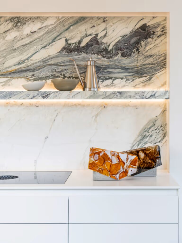 A modern kitchen with white cabinetry, marble-veined splashback, floating lit shelf, and a BROKISGLASS object.
