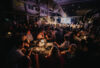Guests dine and converse at candlelit round tables in a chic, industrial venue facing a radiant stage with speakers and screen.