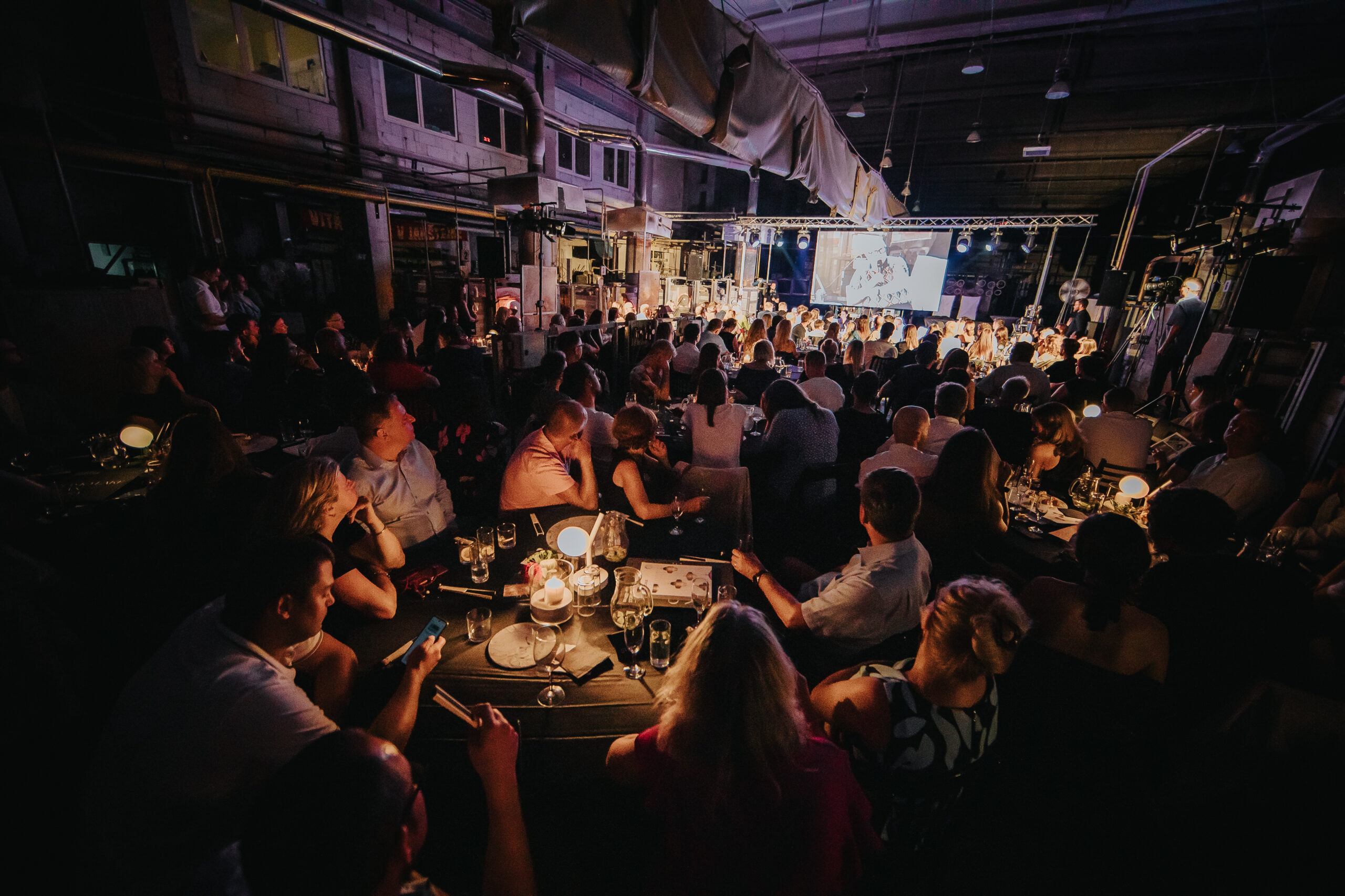 Guests dine and converse at candlelit round tables in a chic, industrial venue facing a radiant stage with speakers and screen.