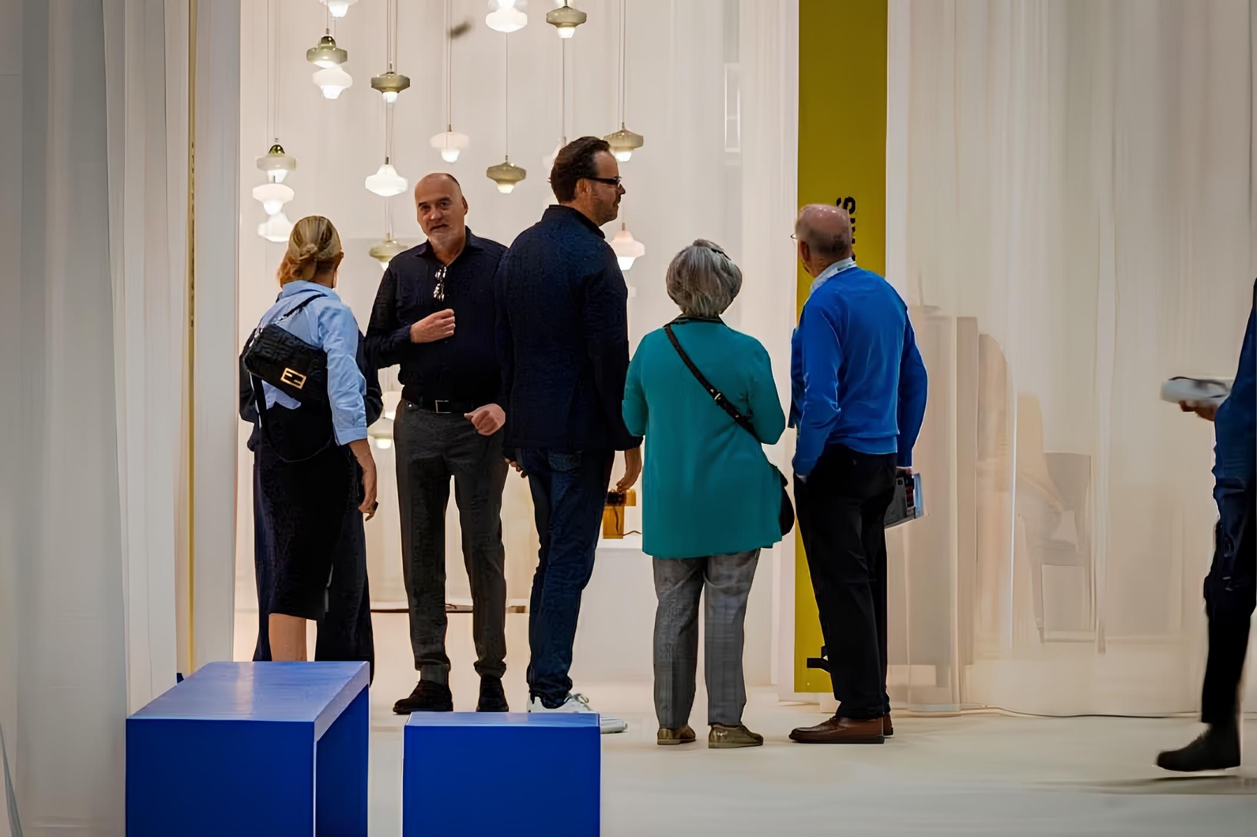 Five adults converse in a modern, sunlit space with flowing white curtains and Brokis Starcloud pendants overhead.