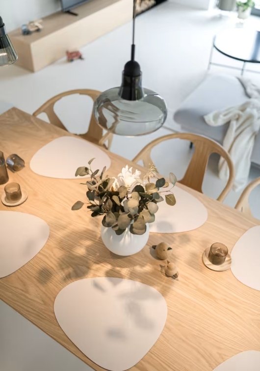 A bright, modern room with a light wooden dining table set with four irregular white placemats, a white vase with green foliage and flowers, candles, wooden bird ornaments, and illuminated by the Shadows Large Pendant overhead.