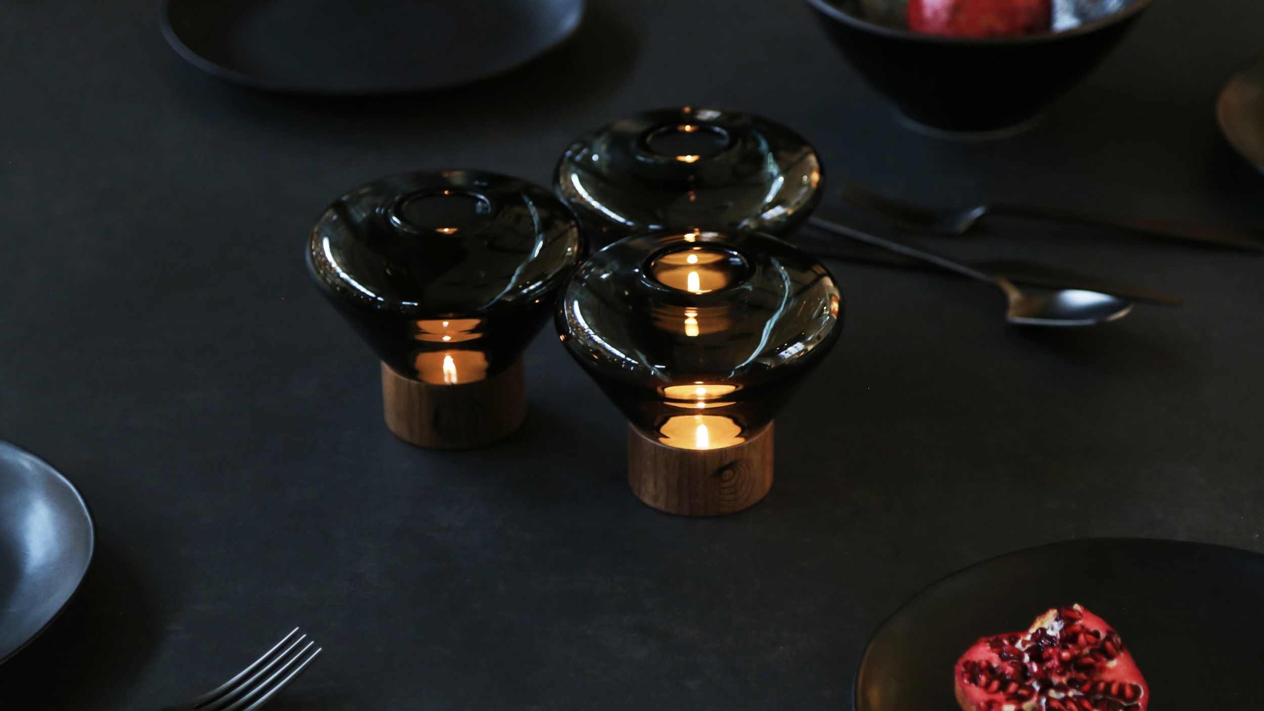 Brokis Muffins Candle holders with glass tops and wooden bases glow on a table, with luxe plates, cutlery, and pomegranate.