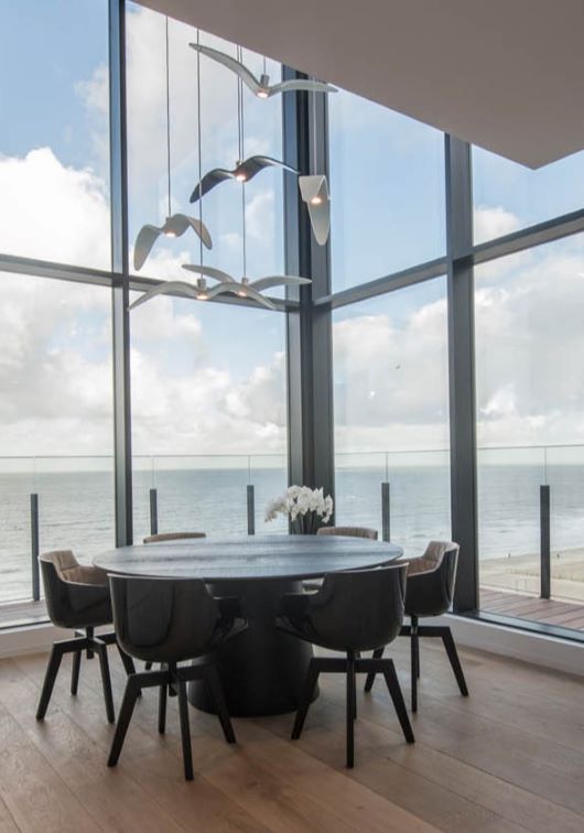A modern dining area with a round table, five chairs, and Brokis Night Birds overlooks the sea through tall windows.
