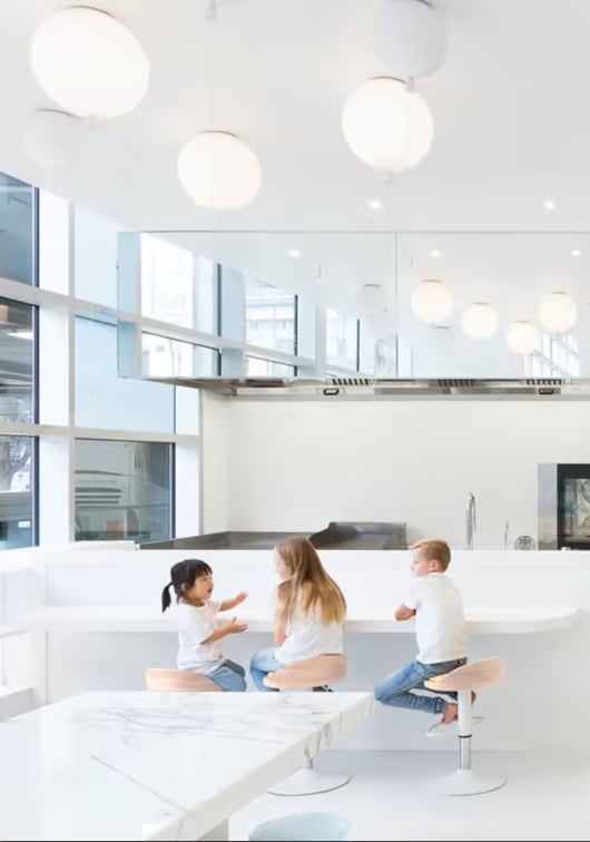 Three children sit at a white table in a minimalist room with Brokis Memory ceiling lights enhancing the airy space.