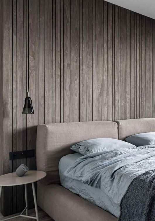 A modern bedroom with a padded taupe headboard, and a Brokis Shadows Narrow pendant above grey wood panelling.