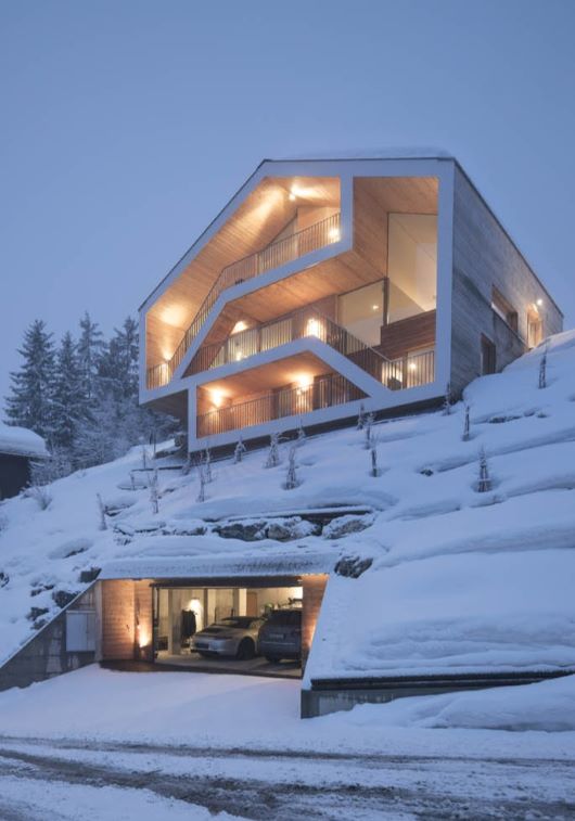 A modern hillside home with expansive balconies glows warmly at dusk, timber façades lit and cars sheltered below amid snow-laden trees.