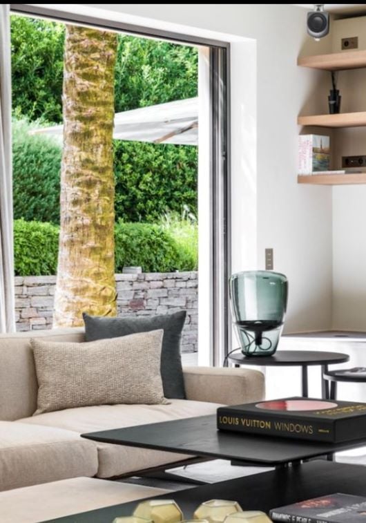 A beige sofa, Brokis Balloons Large above a coffee table with designer books and vase; open doors frame lush garden views.