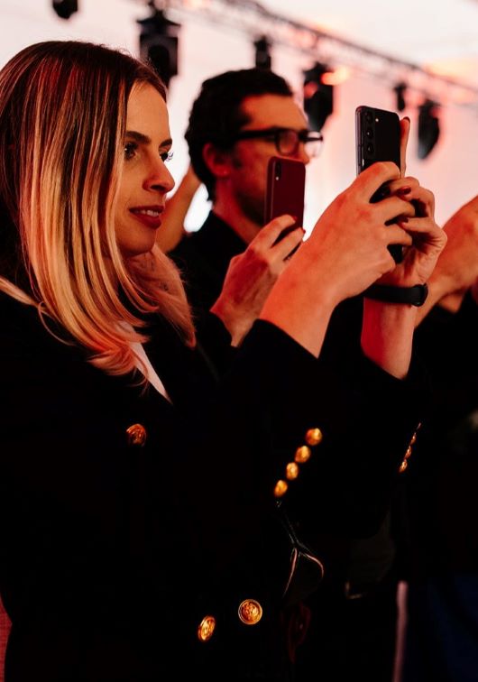 A woman in a black jacket snaps a photo at the Archiproducts Award, warm red tones enhancing the elegant scene.