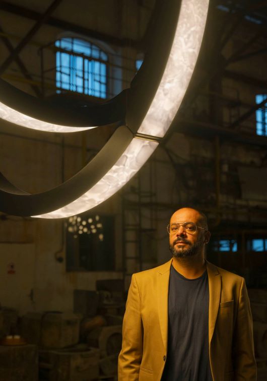 Filippo Mambretti stands beneath the luminous Elo chandelier in an industrial loft with beams and tall daylight windows.