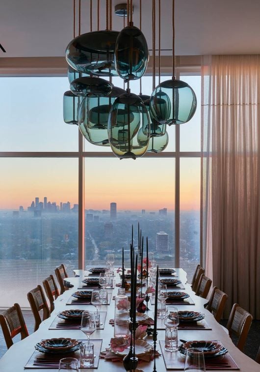 A long dining table faces city sunsets, aglow beneath sculptural, Brokis Knot Pendants and tall black candles.