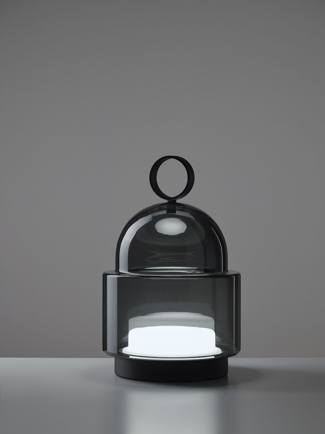 Brokis Dome Nomad Small, with black handle and sculptural smoke grey glass, glows elegantly on a pale surface.