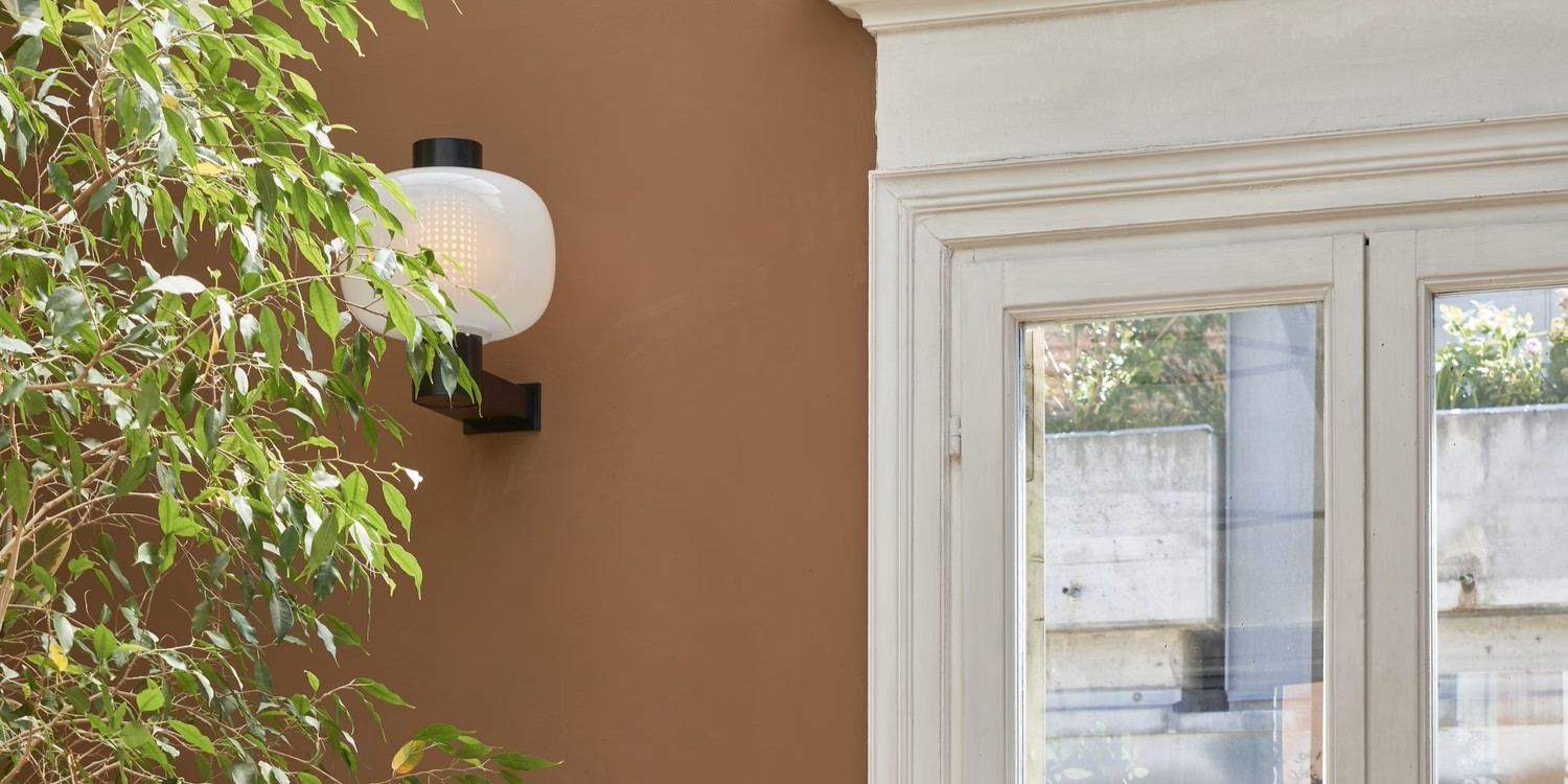 Brokis Bonbori Wall Up above a leafy plant accents a brown wall; nearby, a window frames lush outdoor greenery.