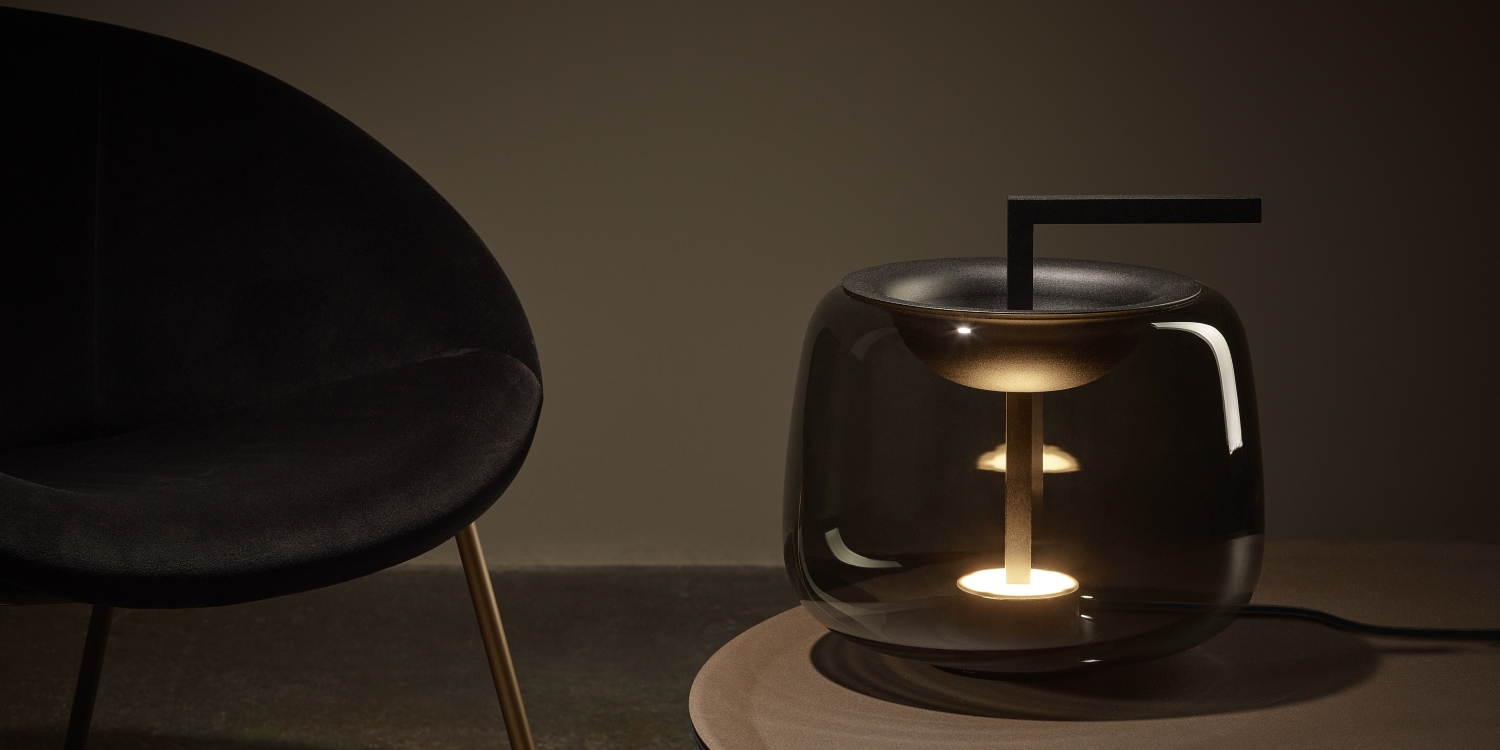 Brokis Double Table lamp glows warmly atop a luxurious table beside a black velvet chair with gold legs.
