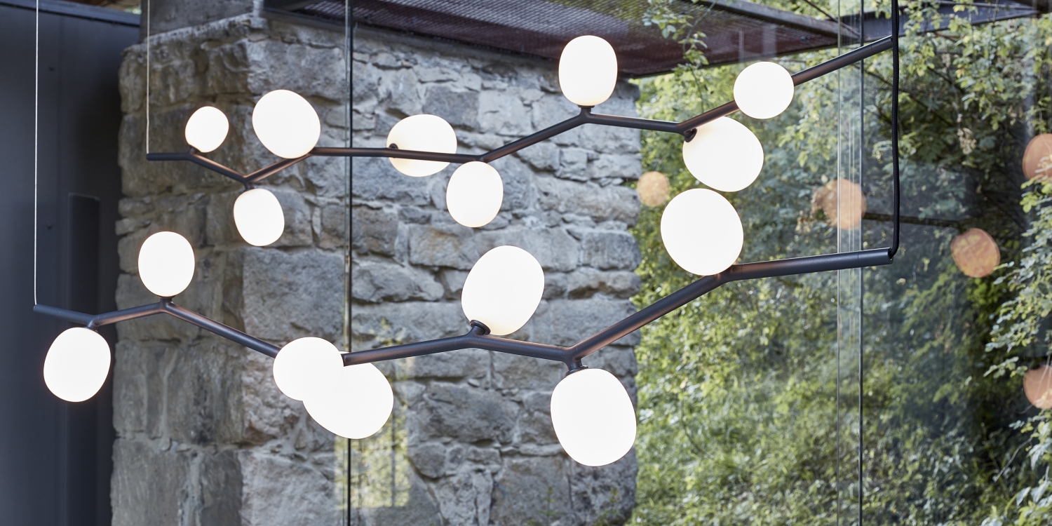 Brokis Ivy Horizontal chandeliers, with hand-blown orbs on sculptural rods, illuminates a glass window framed by lush trees.