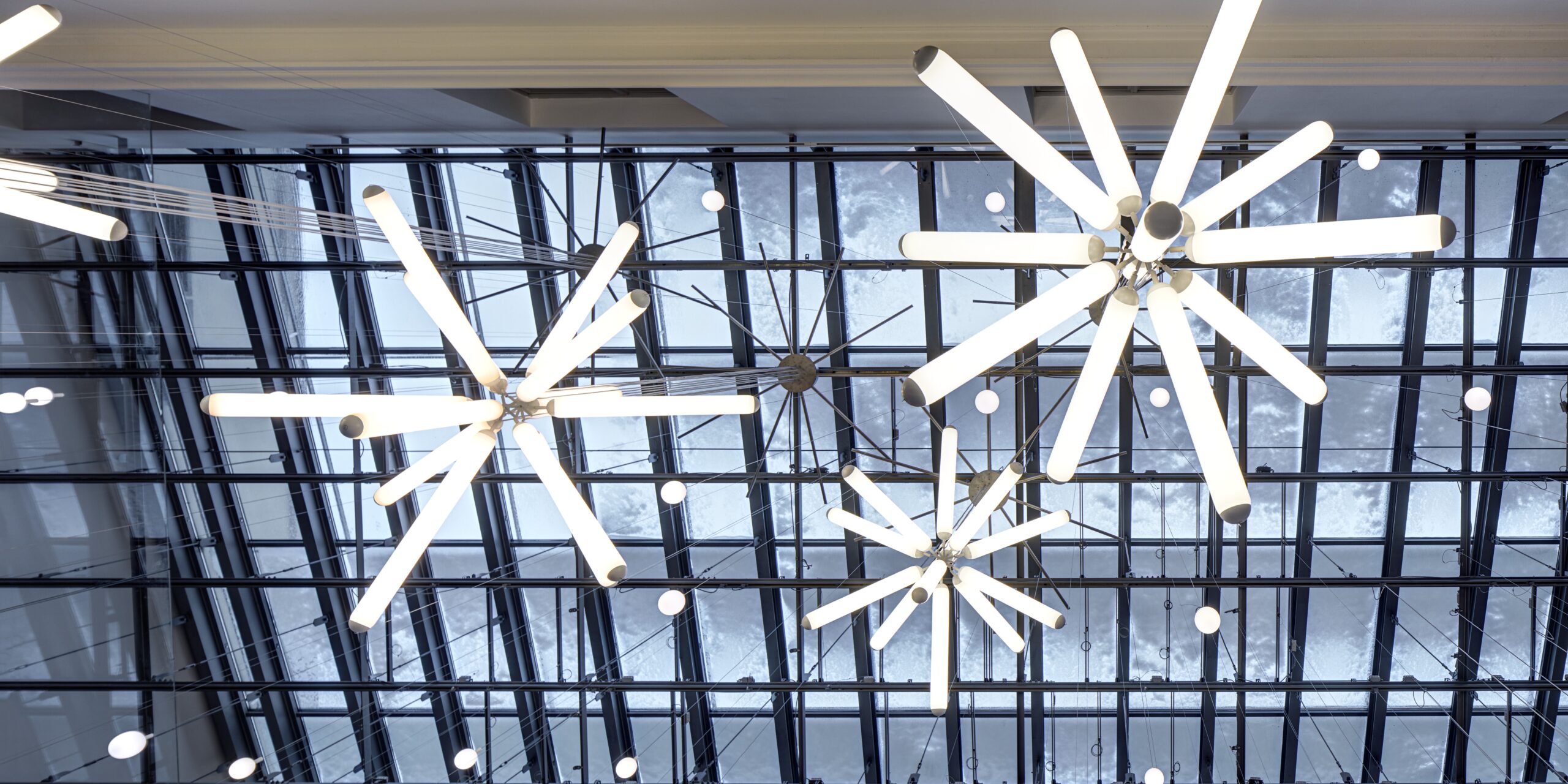 Brokis Puro Sparkle 600 chandeliers glow above a glass grid, their sculptural starbursts reflecting sky and trees beyond.