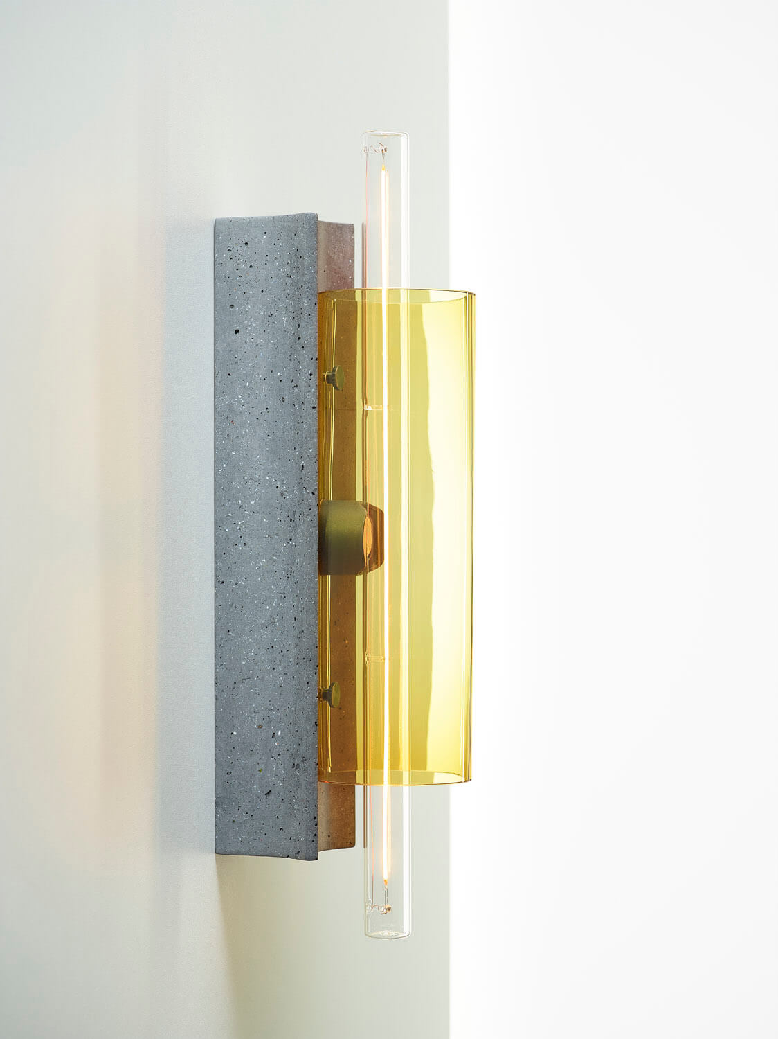 Brokis Overlay Wall M: sculptural concrete base, gold metal detail, amber handblown glass shade, glowing on a white wall.