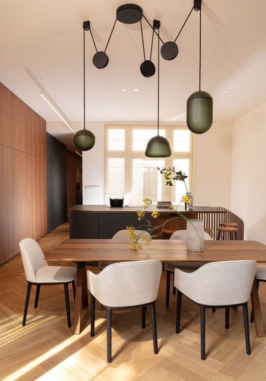 A sunlit dining room with a wooden table, beige chairs, and Brokis Planets 3 Set pendants highlights elegant minimalism.