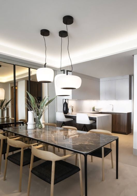 A modern dining room with a glass table, woven chairs, and handblown Brokis Double pendants above; lush greenery accents.