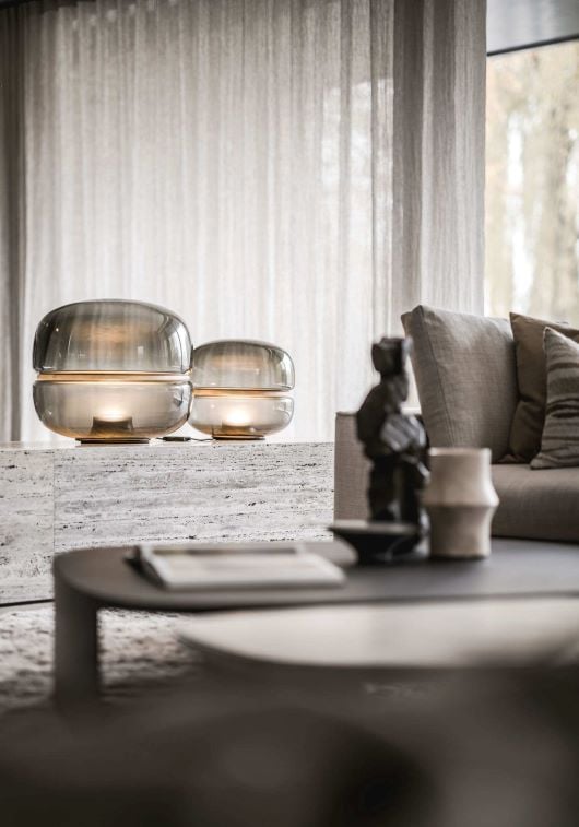 A modern living room with a beige sofa, sculptural Macaron glass lamps on stone, and luxe accents beneath sheer curtains.