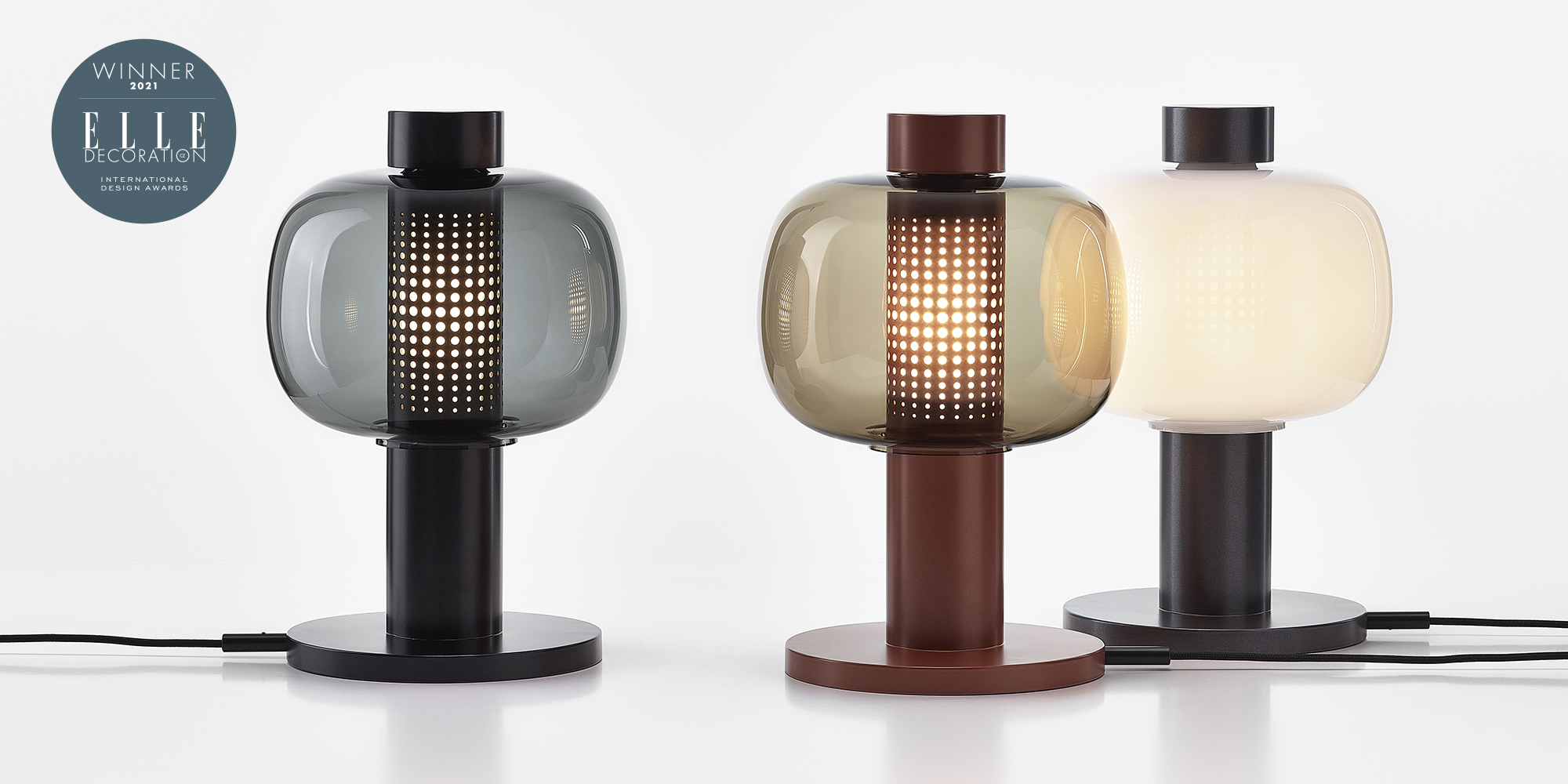 Three Brokis Bonbori table lamps with hand-blown glass shades in smoky grey, amber, and white on cylindrical bases.