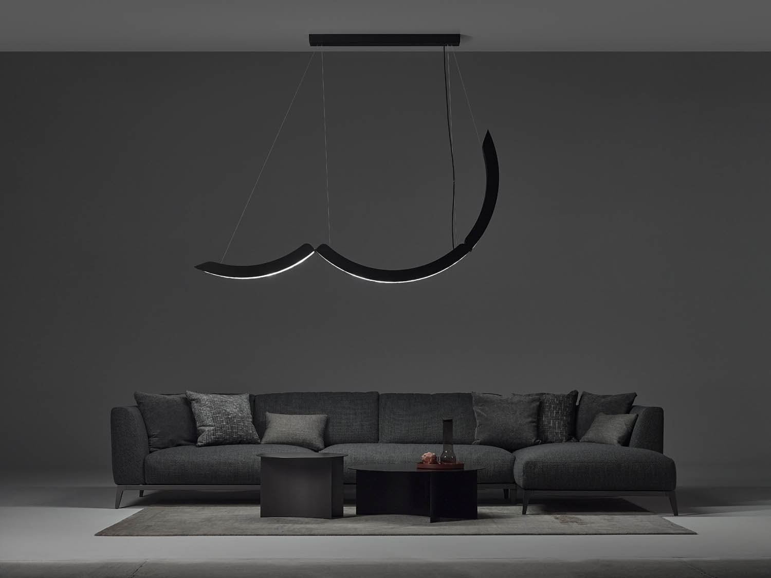 A modern living room with a dark grey sectional, minimalist rug, twin black coffee tables, and a Brokis Elo Chandelier above.