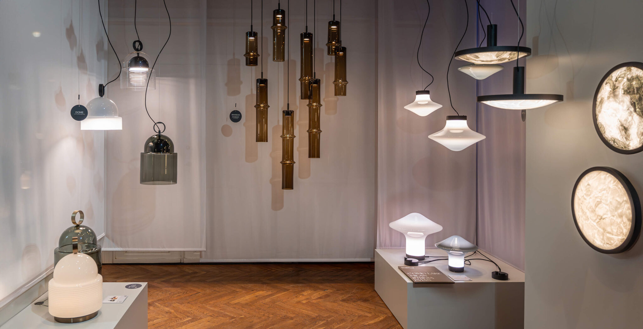 A lighting showroom features sculptural, hand-blown Brokis pendant, wall, and table lamps aglow on sleek white plinths.