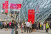 A stylish crowd with luggage gathers outside a geometric glass building under bold red Salone del Mobile Milano signs.