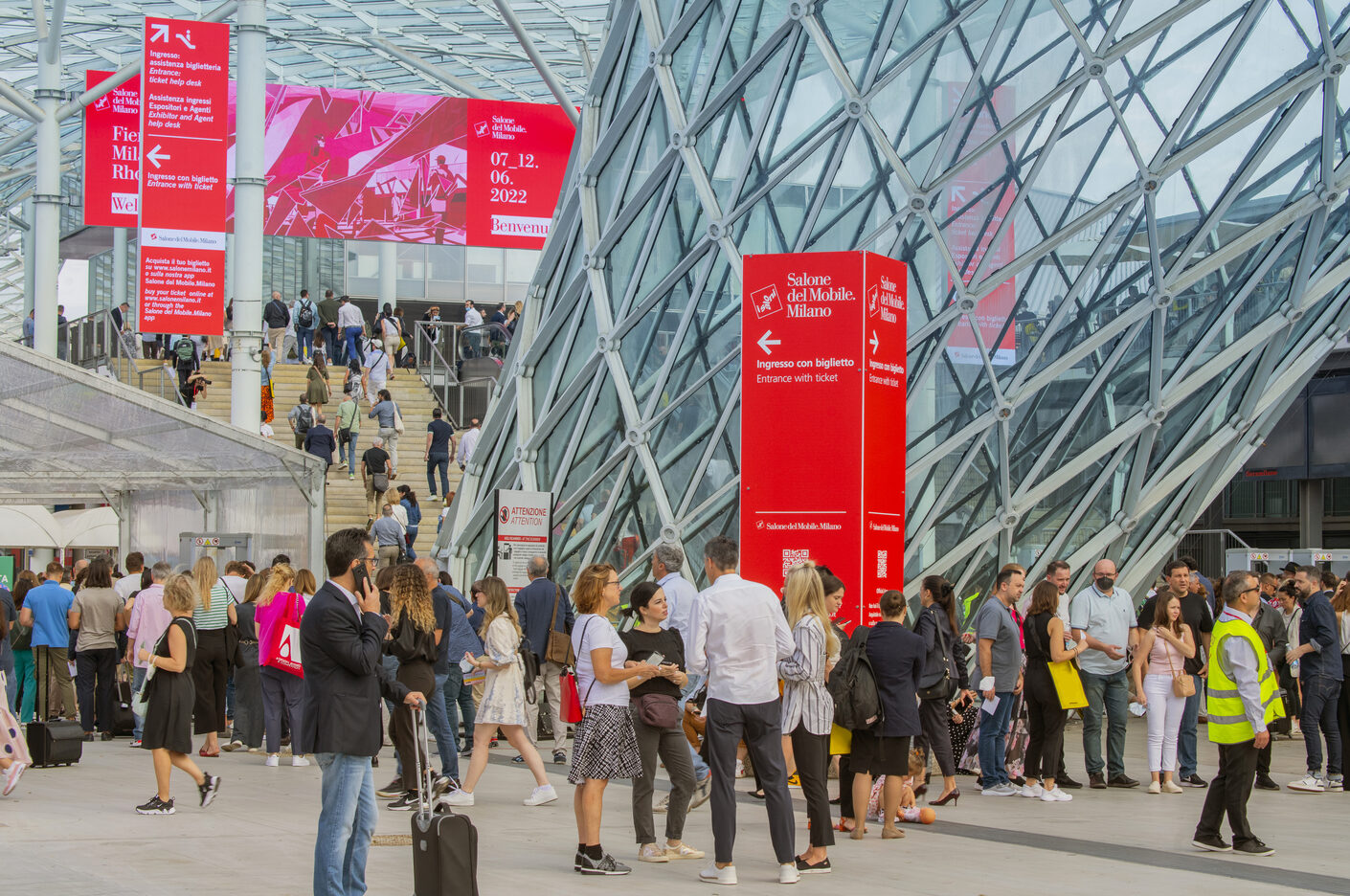 A stylish crowd with luggage gathers outside a geometric glass building under bold red Salone del Mobile Milano signs.