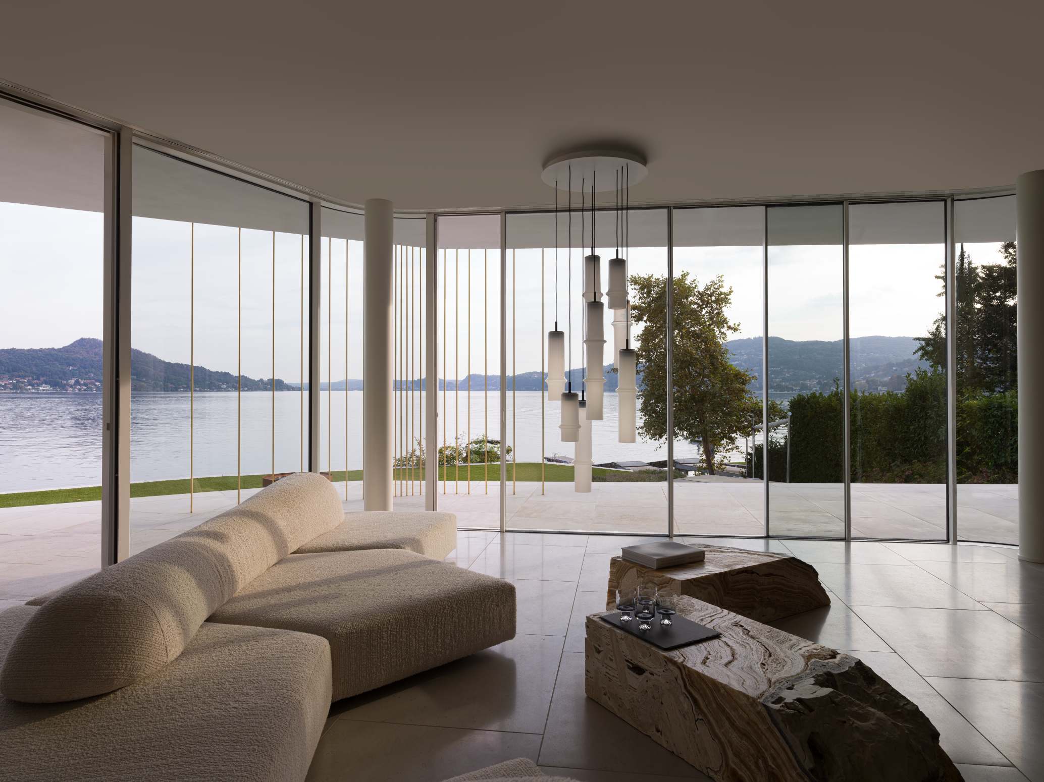 A modern living room overlooks a lake; a curvy cream sofa and Brokis Bamboo Forest lights add refined serenity.