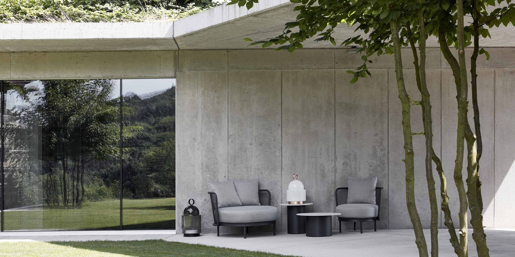 A modern patio with two grey armchairs, Brokis Dome Nomads, and chic accents set against lush foliage and concrete.