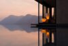 Elegant modern home with glass walls, Brokis Muffins lamps, and chair reflecting in an infinity pool at sunset.