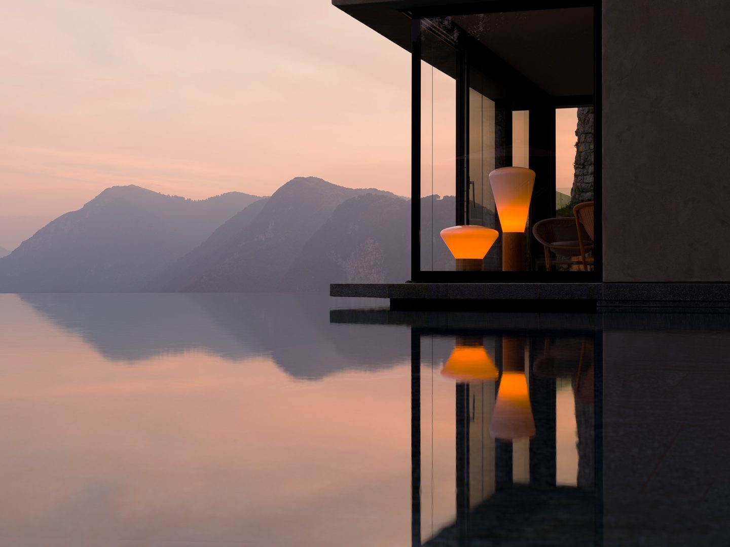 Elegant modern home with glass walls, Brokis Muffins lamps, and chair reflecting in an infinity pool at sunset.