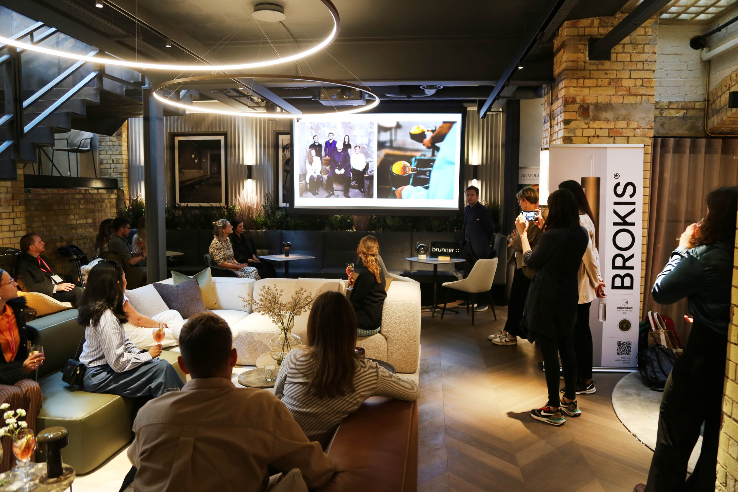 Attendees gather in a chic, exposed-brick lounge with designer furnishings, captivated by a stylishly projected presentation.