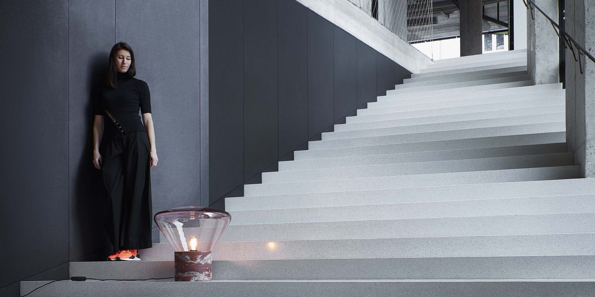 Lucie Koldva stands by a dark wall and modern stairs, illuminated by a Brokis Muffins Wood 02 floor lamp.