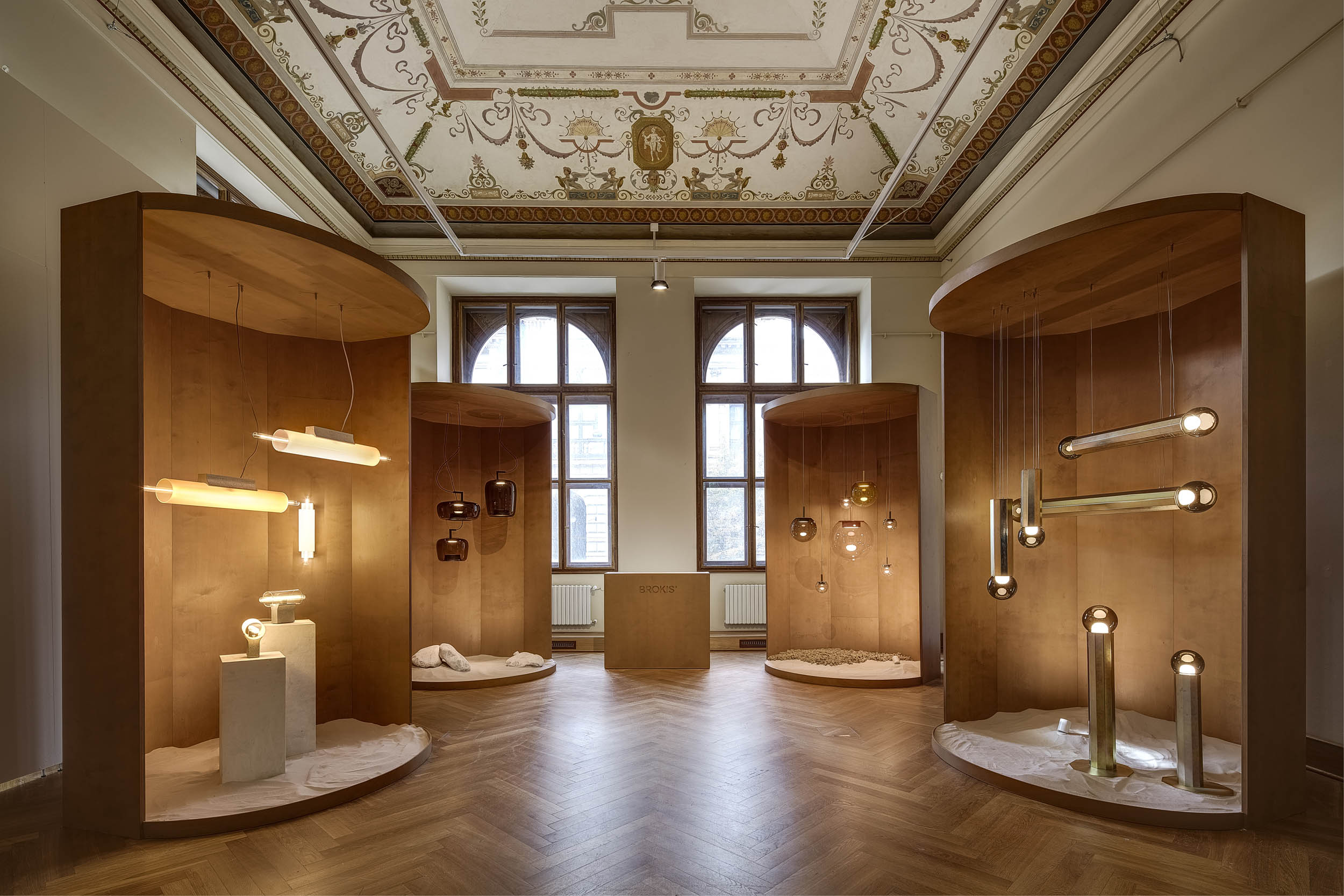 An elegant room with ornate ceiling and arched windows features hand-blown Brokis lights in warm wooden alcoves.