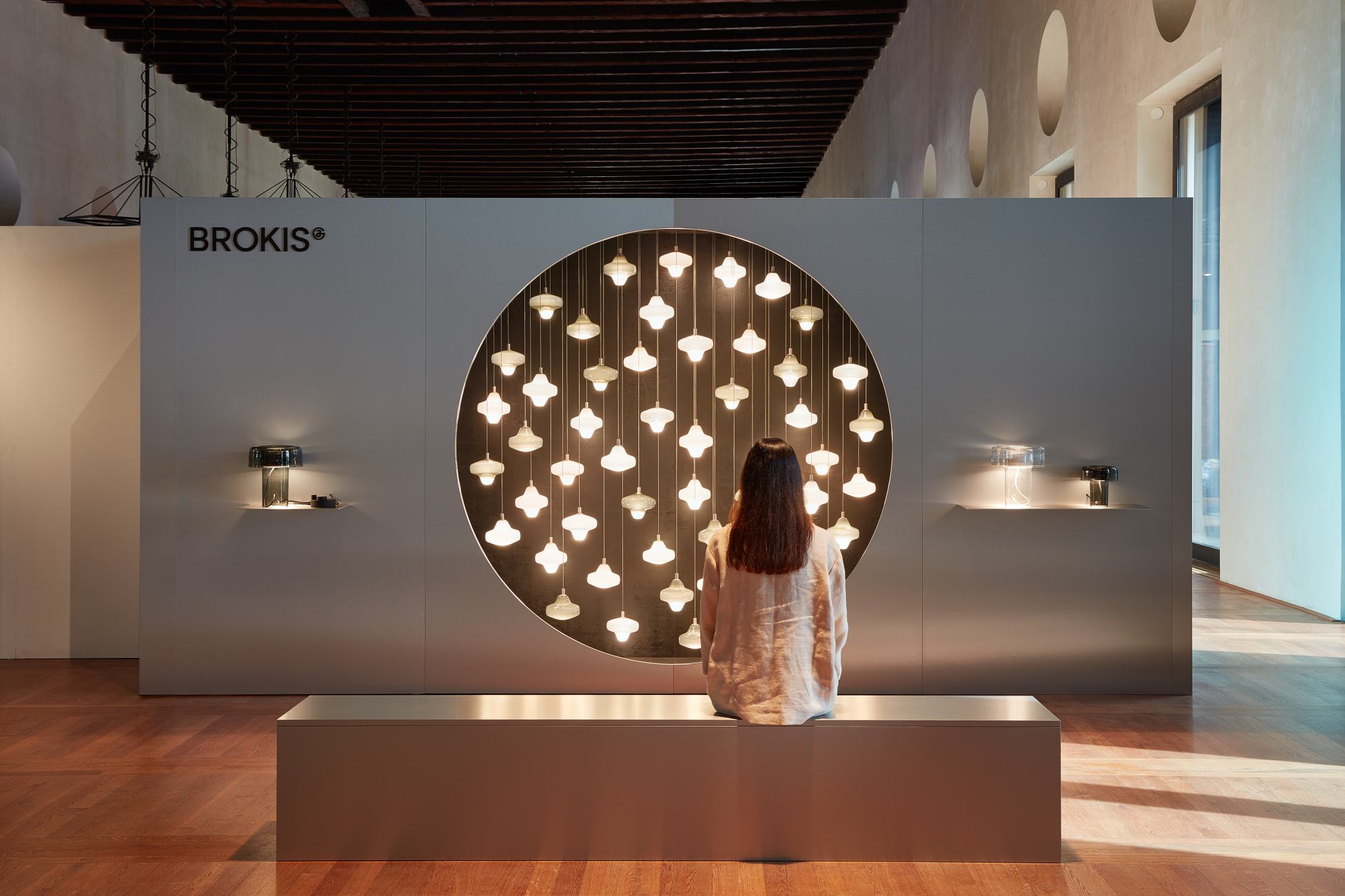 A woman sits on a grey bench, admiring glowing Brokis Starcloud lights set in a luxurious modern art display.