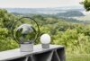 Brokis Sfera Portable lamps with metal bases grace a concrete table, lush greenery beyond under a moody sky.