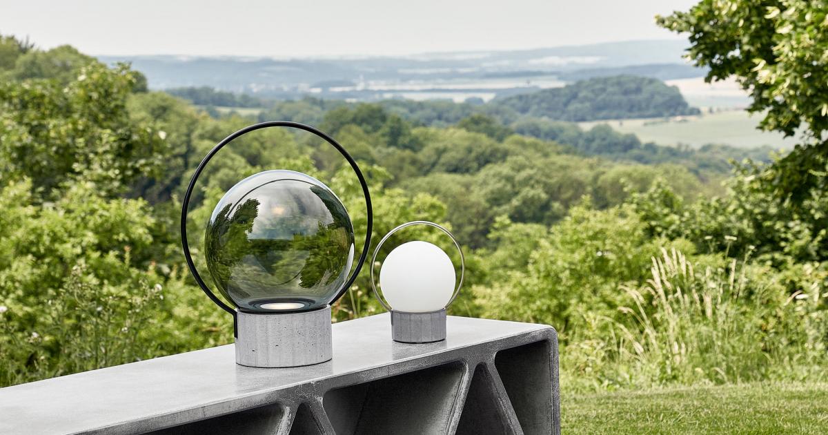 Brokis Sfera Portable lamps with metal bases grace a concrete table, lush greenery beyond under a moody sky.
