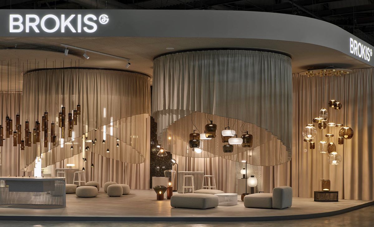 Luxurious stand showcasing sculptural handblown Brokis lights, plush seating, curved beige drapes, and glowing logo.