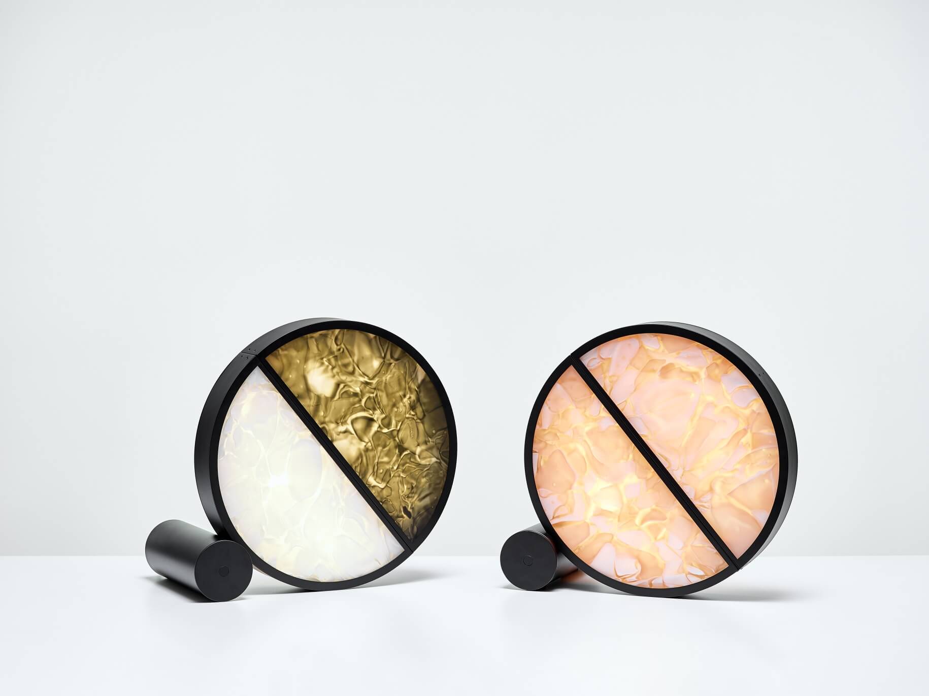 Two Brokis Geometric Table lamps in gold-white and pink-yellow rest at an angle on a white surface; black bases gleam.