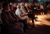A diverse group, elegantly dressed in dark tones, sits closely at an indoor event, bathed in luxurious warm stage lighting.