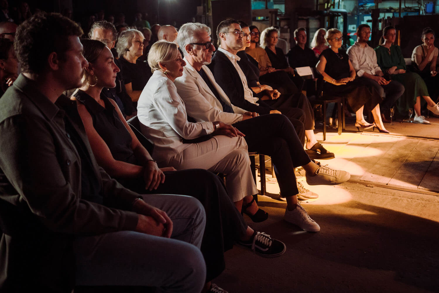 A diverse group, elegantly dressed in dark tones, sits closely at an indoor event, bathed in luxurious warm stage lighting.