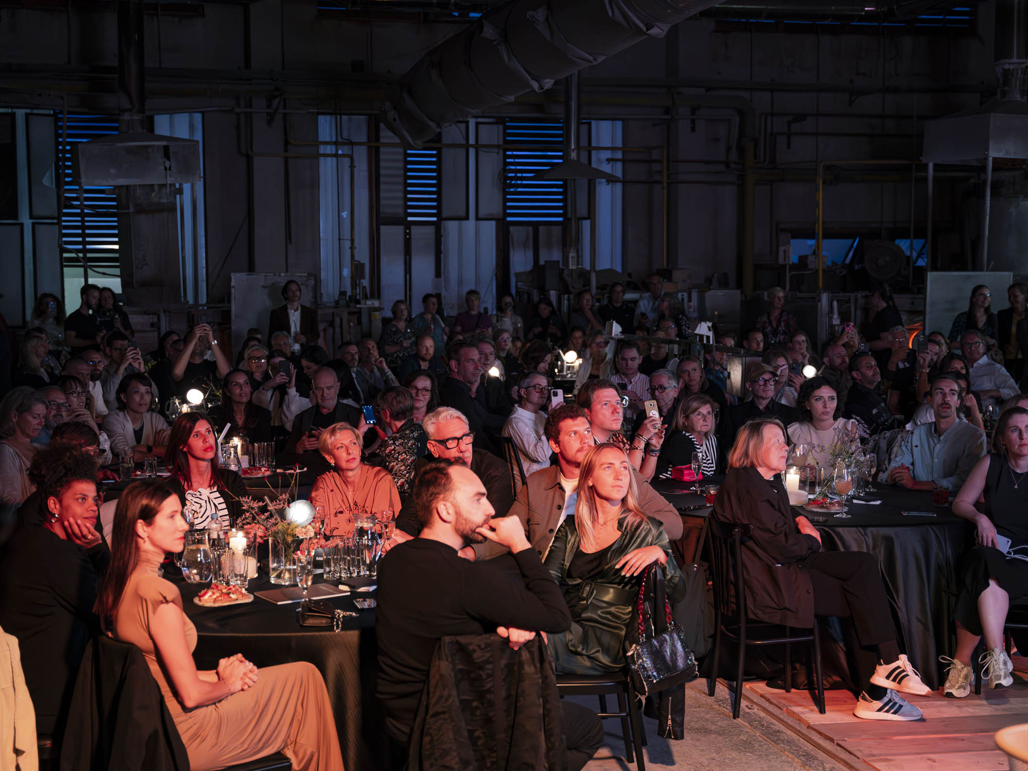 A sophisticated crowd sits at round tables in a moody industrial venue, faces aglow as Brokis Ivy Battery lamps cast soft light.