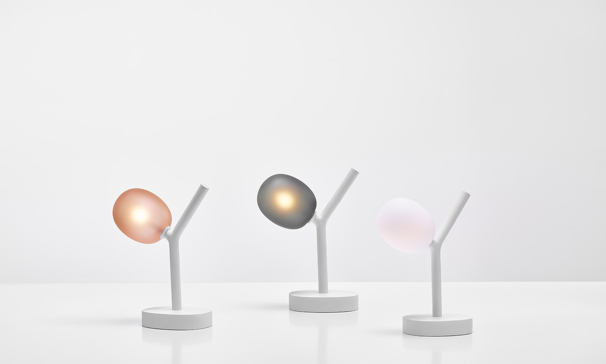 Three Brokis Ivy Battery table lamps with branch-like white bases and frosted glass glow in pink, grey, and white.