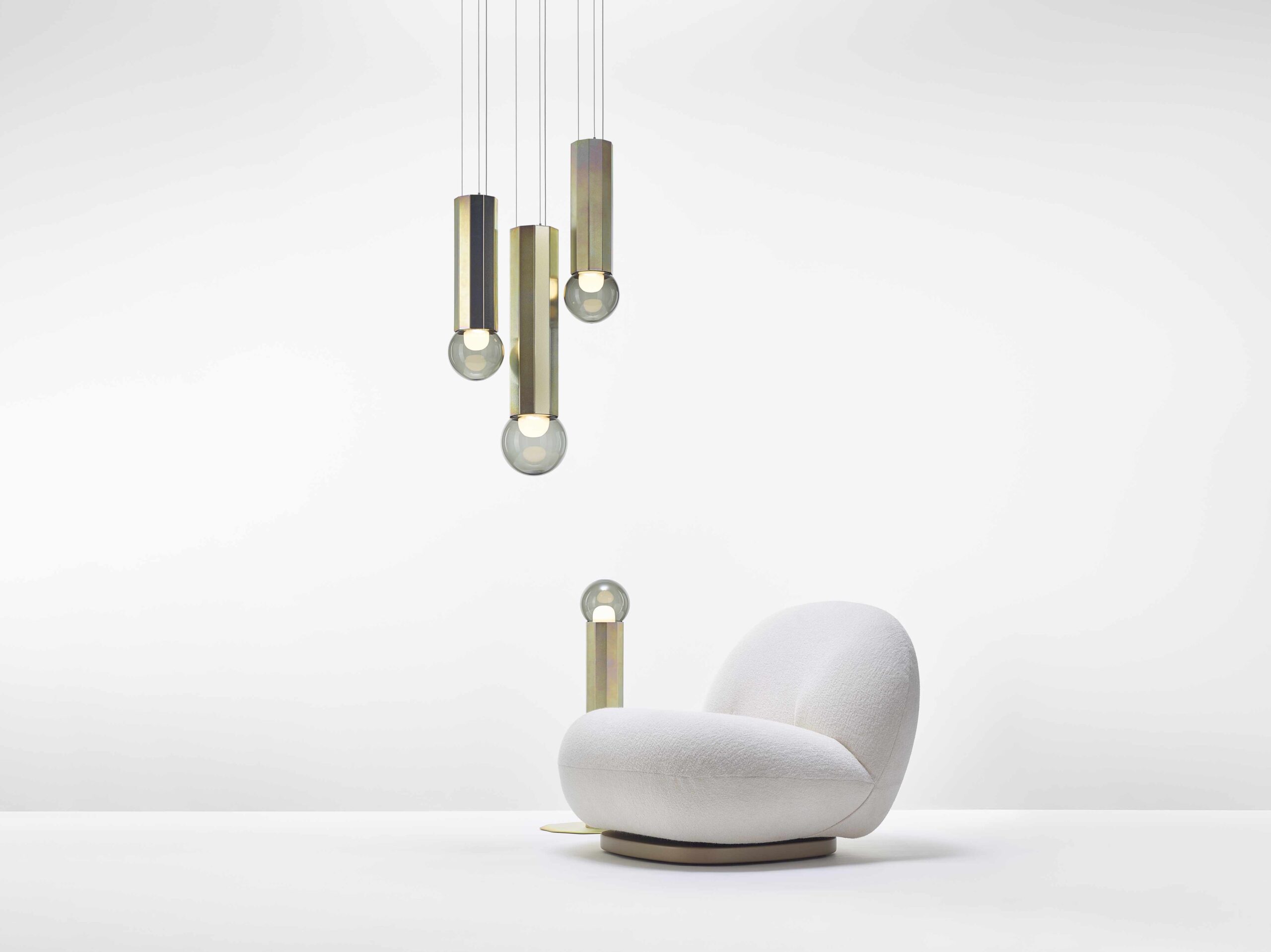 A sculptural white lounge chair on a pristine floor beneath three Brokis Prisma pendants, set against a crisp backdrop.