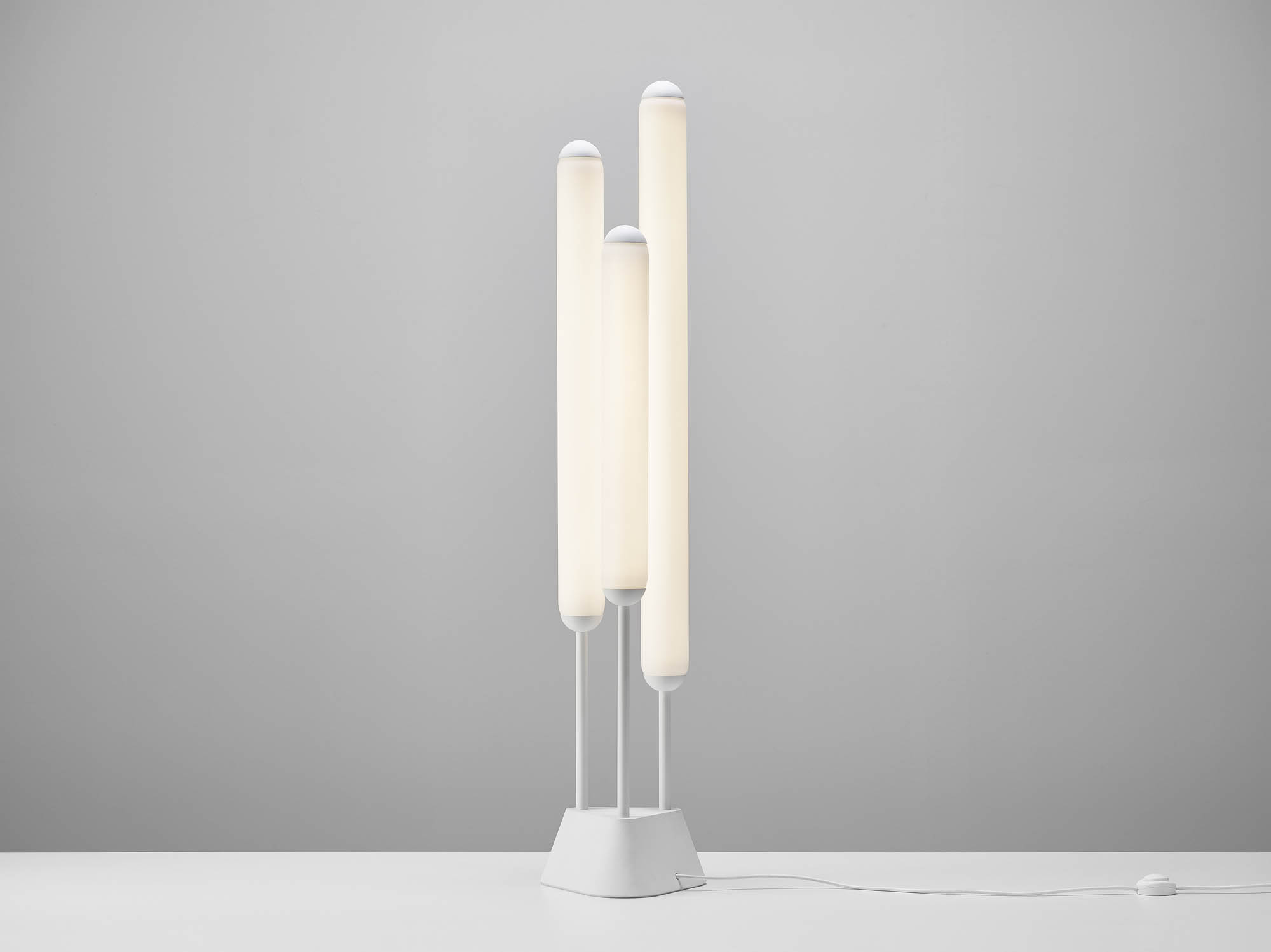 A Brokis Puro floor lamp with three hand-blown, frosted crystal tubes of varying heights glows atop an angular white base.